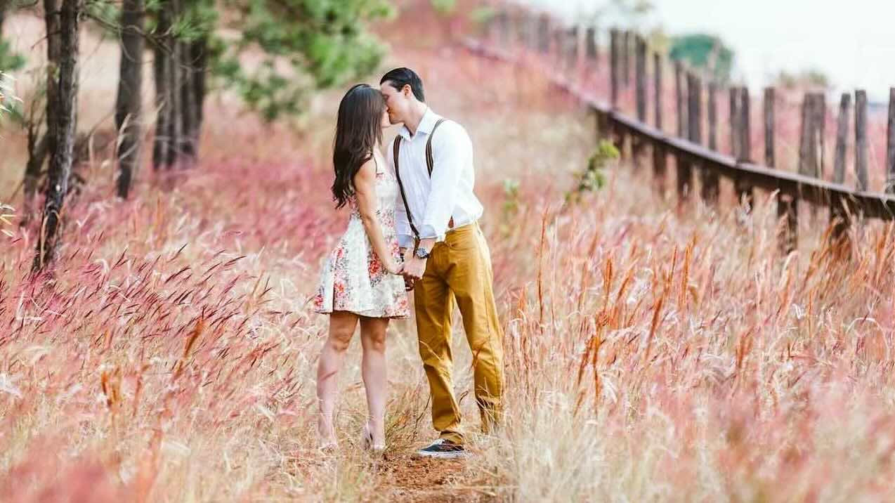 A man and woman wearing country clothes are standing in a field of pink wildflowers kissing.