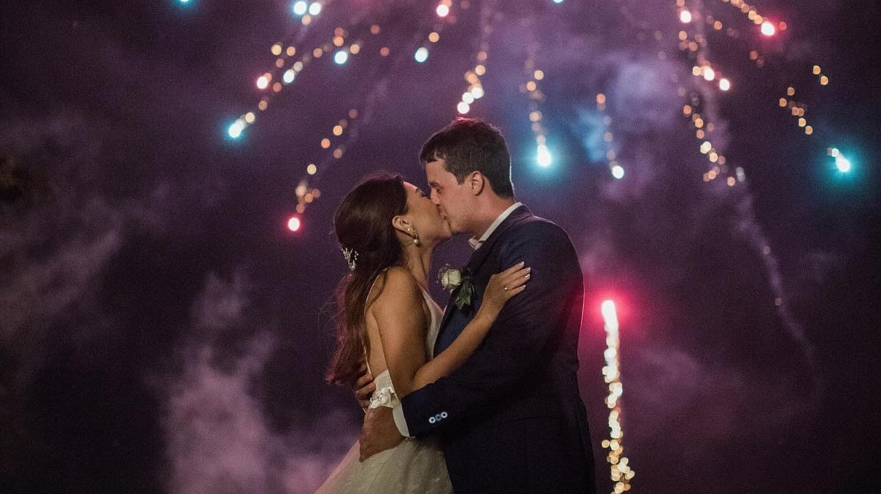 Bride and groom kissing under a night sky with fireworks.