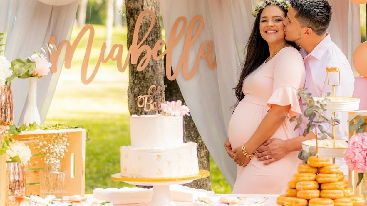 Pregnant woman in pink dress is hugged by her husband, while being surrounded by baby shower decorations.