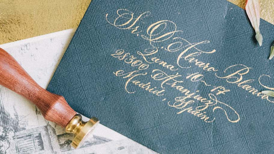 Blue wedding invitation envelope with elegant gold writing on it. The envelope is sitting on a gold tray with flower petals