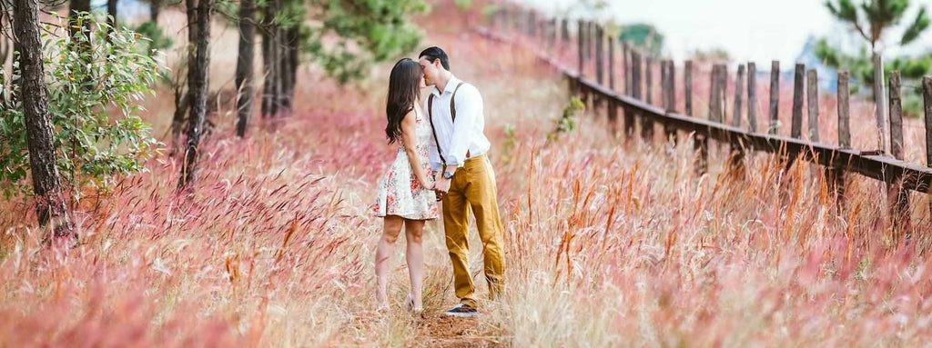 A man and woman wearing country clothes are standing in a field of pink wildflowers kissing. 
