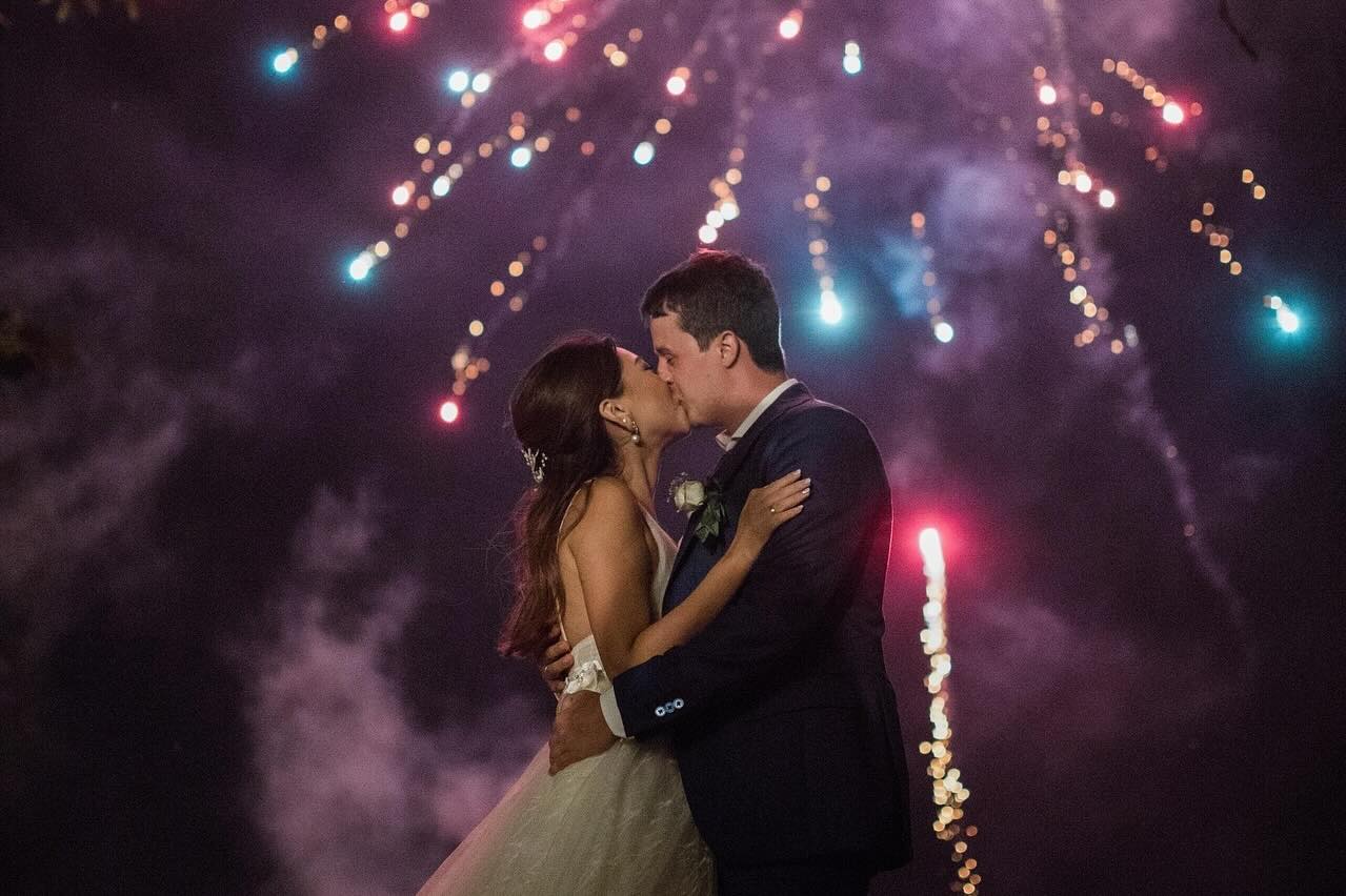 Bride and groom kissing under a night sky with fireworks.