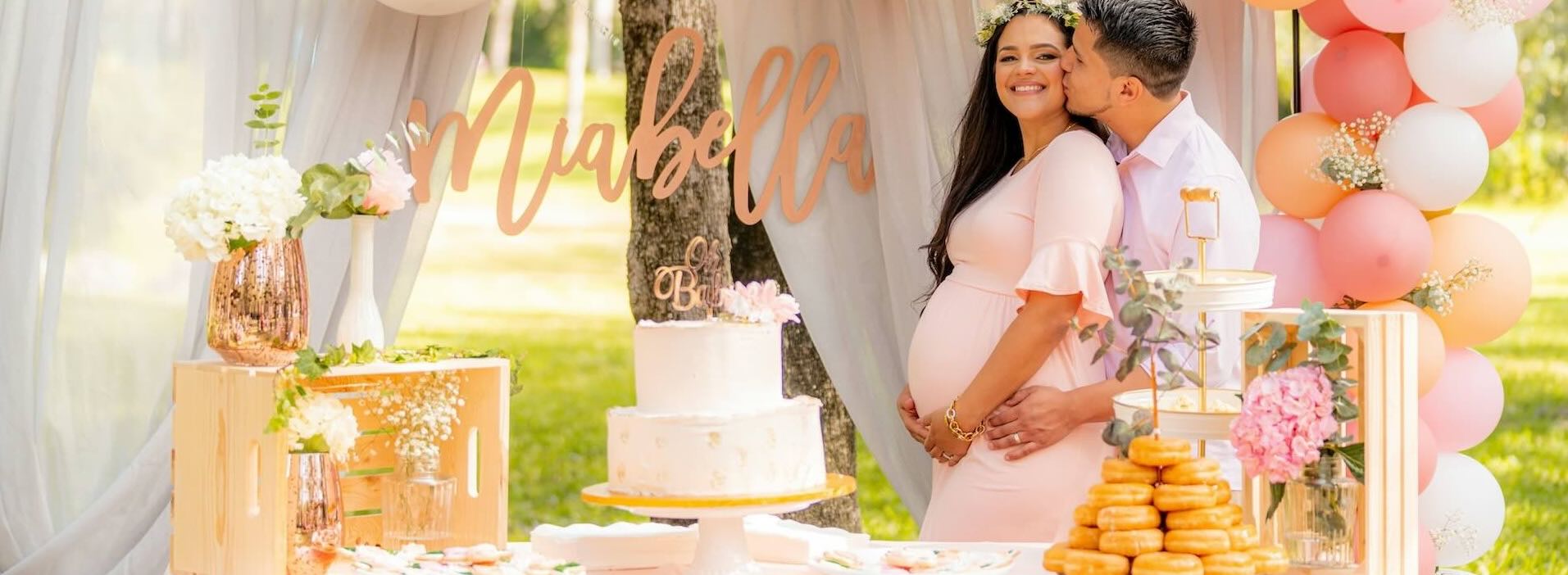 Pregnant woman in pink dress is hugged by her husband, while being surrounded by baby shower decorations.