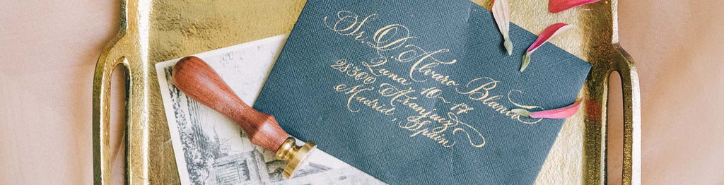 Blue wedding invitation envelope with elegant gold writing on it. The envelope is sitting on a gold tray with flower petals